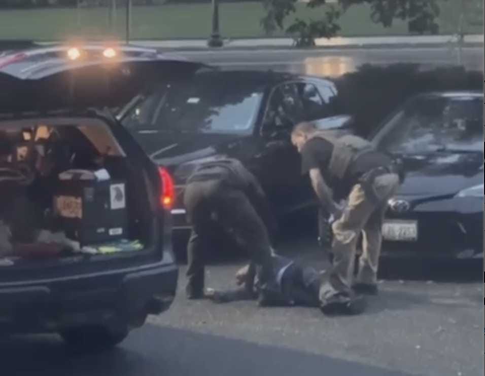 Two officers in tactical gear restrain a person on the ground between parked cars in a parking lot with a car trunk open nearby