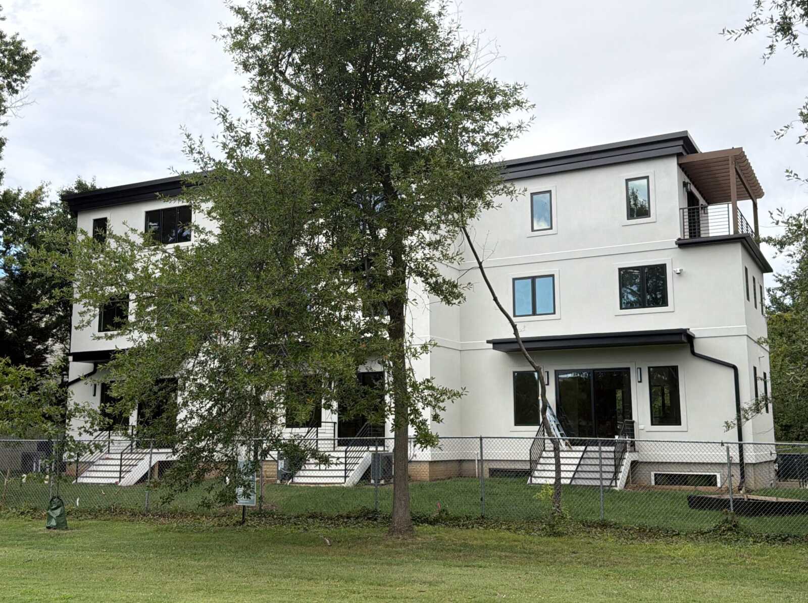 Modern white multi-story building with large windows, a balcony on the right, and a metal staircase, set behind a chain-link fence and trees on a grassy lot