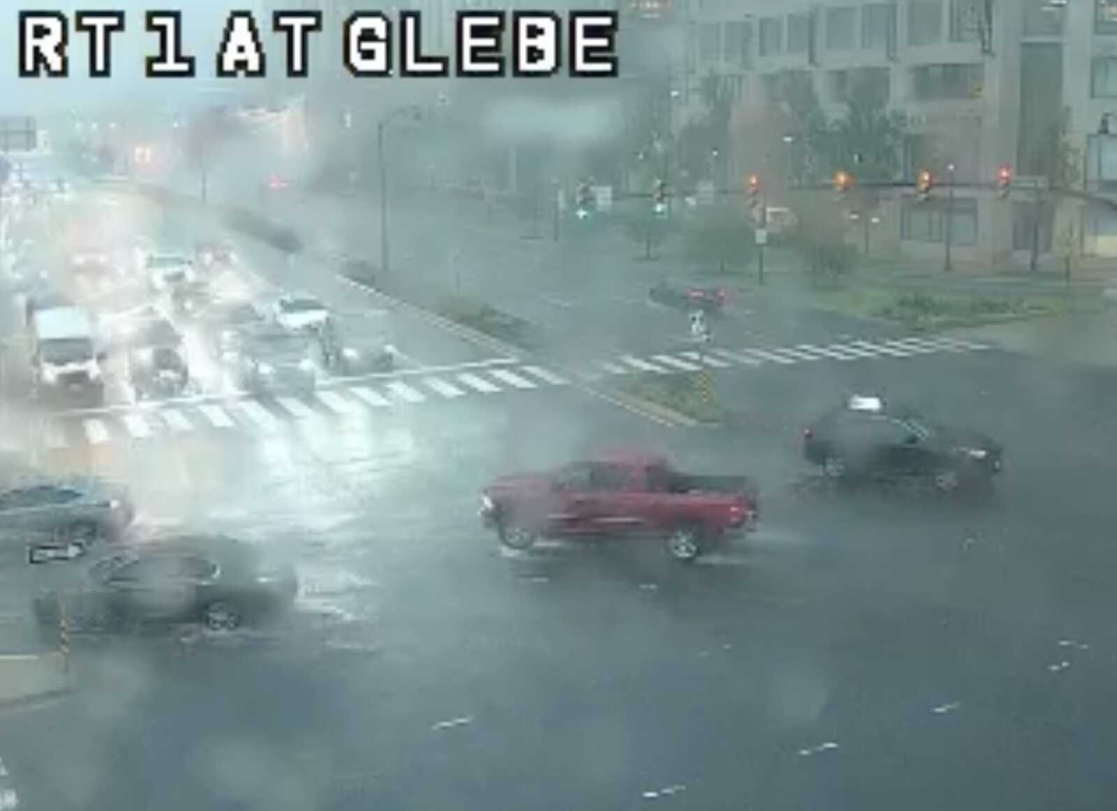Rainy city intersection with several cars and a crosswalk; overlay text reads 'RT1 AT GLEBE' at the top left.