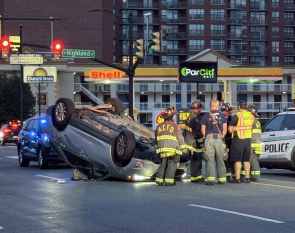 Overturned sedan in the middle of an intersection as firefighters and police stand nearby at dusk, assessing the scene.