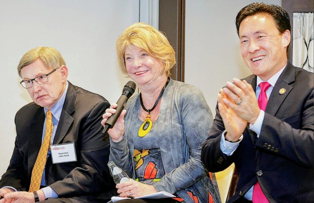Three panelists at a conference: a woman in the center speaks into a handheld microphone while two men in suits listen—one on the left looks serious, the other on the right applauds.