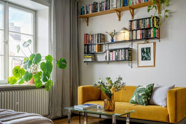 Cozy living room with a mustard-yellow sofa, glass coffee table, and wall-mounted bookshelves filled with books.
