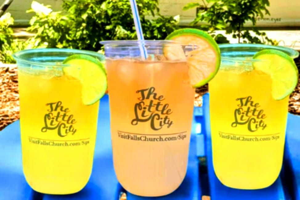 Specialized drink glasses are part of the Little City Sips marketing effort (via City of Falls Church) | ARLnow Three colorful iced drinks in clear cups with lime wedges on a blue table outdoors; each cup bears 'The Little City' logo and a Falls Church SIP URL.
