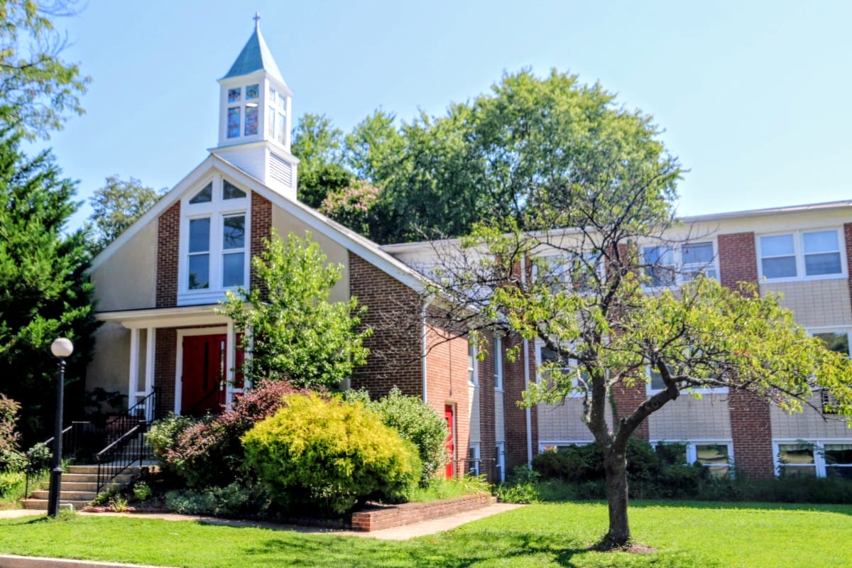 Brick church with a white steeple and red doors, set among manicured shrubs and trees on a sunny day.