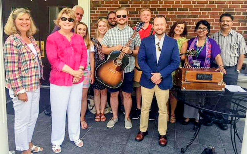 Group of ten adults posing together on a patio; a man with an acoustic guitar stands in the center among smiling colleagues.