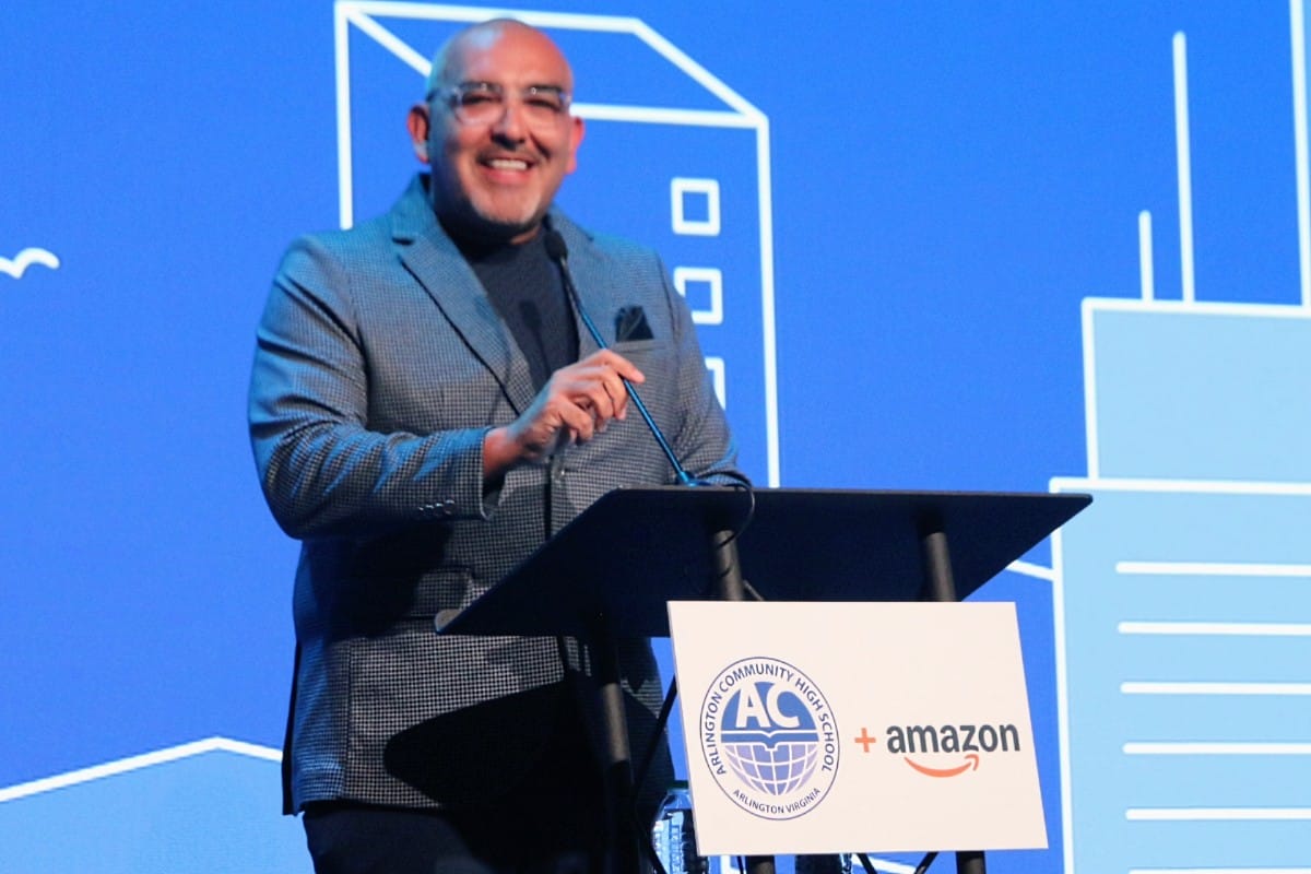 Smiling man in a gray blazer speaks at a podium during a conference, with a blue backdrop and a sign showing AC High Schools and Amazon logos at the lectern