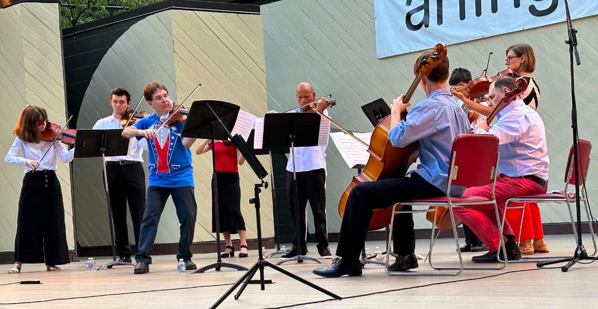 The National Chamber Ensemble performs at Lubber Run Amphitheater (photo by Steven Fleshman) | ARLnow A string ensemble performs on an outdoor stage, with violinists standing and a cellist seated, with music stands and sheets in view.