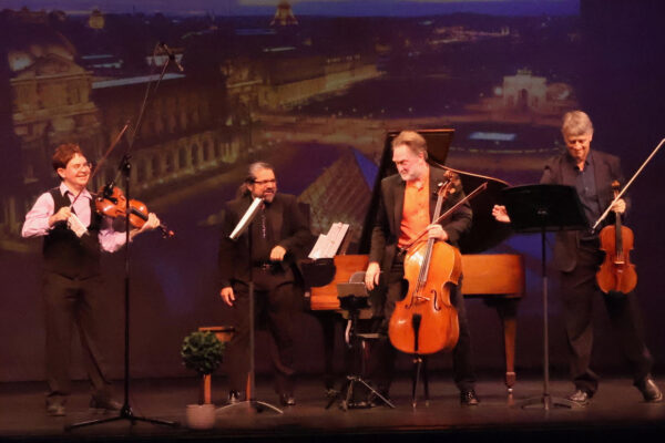 A quartet performing on stage: violinists/flutists and a cellist with a pianist, in front of a cityscape backdrop.