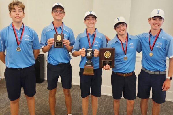 Five young athletes in matching blue shirts and white caps pose with medals and trophies after a win? holding a cup and plaques in a hallway.