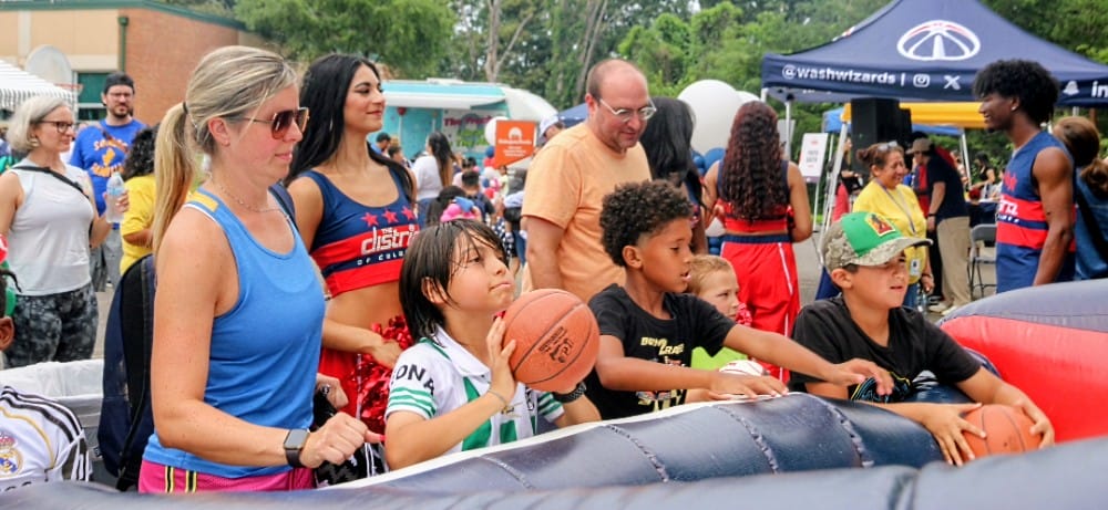 Children and adults at an outdoor festival, guiding basketballs toward a inflatable obstacle course at a sunny day.
