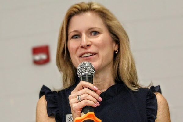 Woman speaking into a handheld microphone, smiling, wearing a dark sleeveless top or blouse in a talk setting.