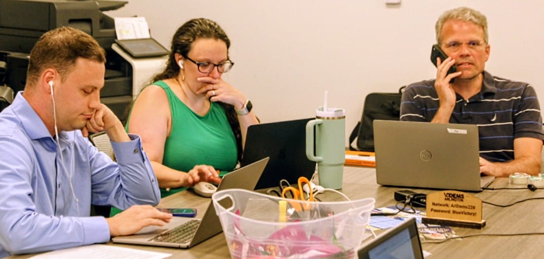 Three professionals sit at a conference table with laptops: left man wearing a blue shirt using a laptop and earbuds, center woman in green examining a tablet, right man on a phone with a Dell laptop nearby.