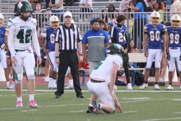 On-field football scene with a kneeling player in white and green, a referee nearby, and opposing players in blue and gold watching from the sideline.