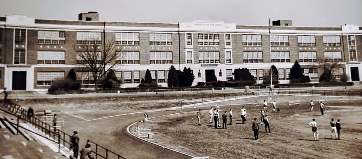 Black-and-white photo of a brick school building with a dirt field and groups of students on recess.