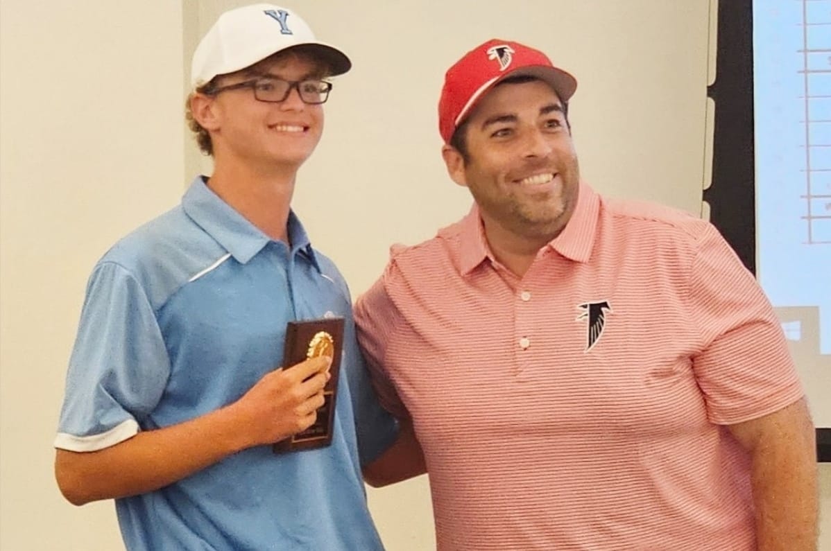Two men pose for a photo at an awards event: a young golfer in a light blue polo and white cap holding a plaque, smiling beside another man in a pink striped polo and red cap with a Falcons logo appears proud.]