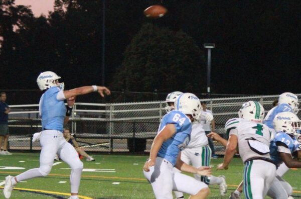 Quarterback in light blue jersey throws a pass as teammates and defenders watch the ball in the air on a nighttime football field.