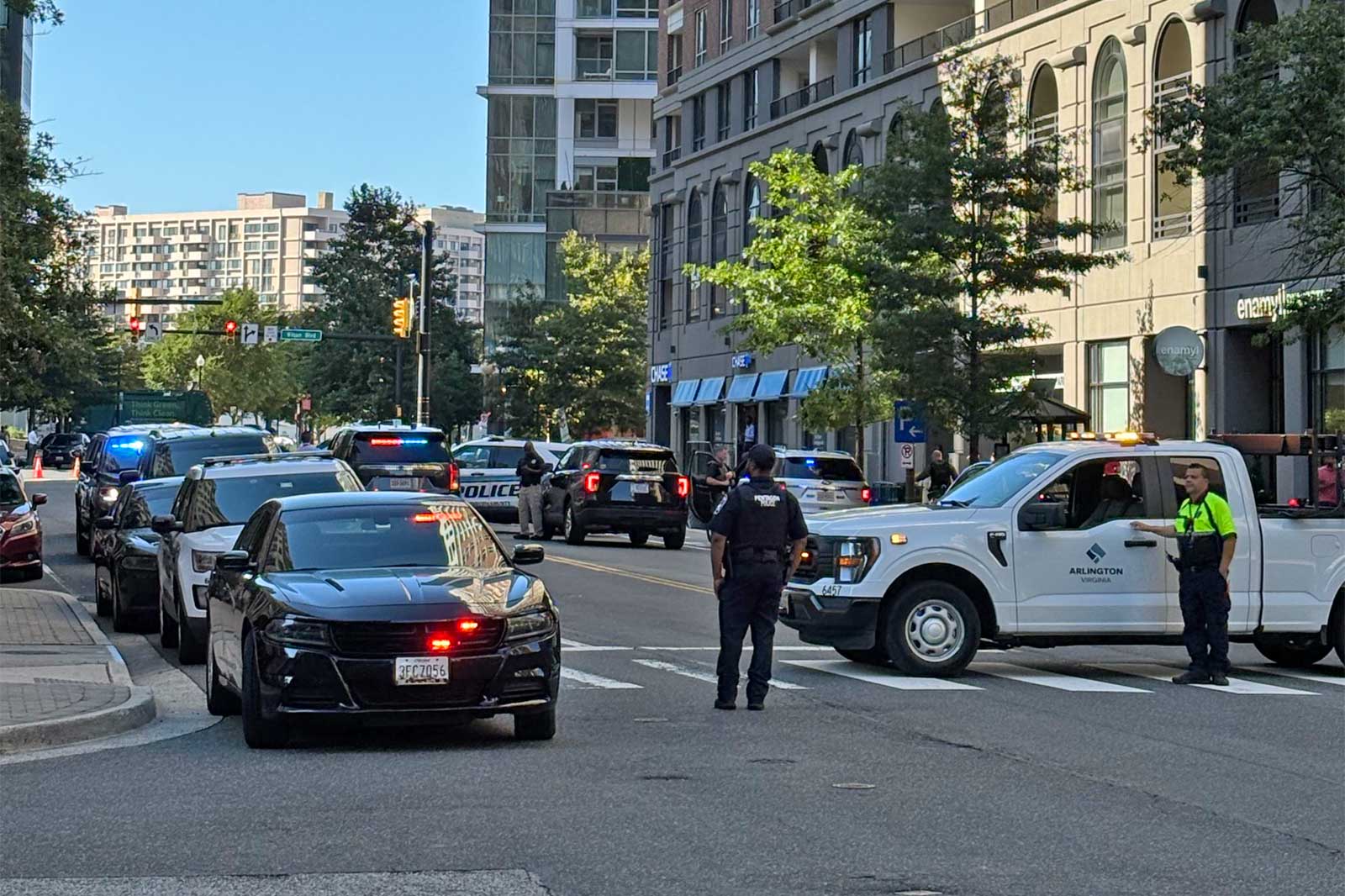 Police cars with flashing lights block a City street; officers stand near a white Arlington, Virginia vehicle on a busy urban intersection.