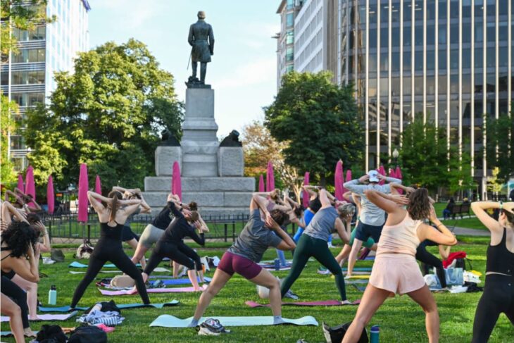 Group of people practicing yoga on mats in a city park, with a statue on a pedestal and modern buildings in the background nudging the skyline.