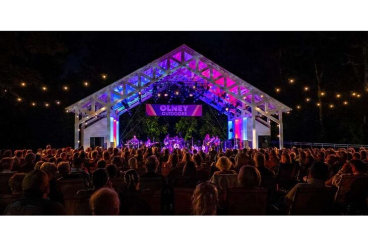 Outdoor night concert with a brightly lit open-air stage and a large seated audience; banner reads 'OLNEY OUTDOORS' at center stage.