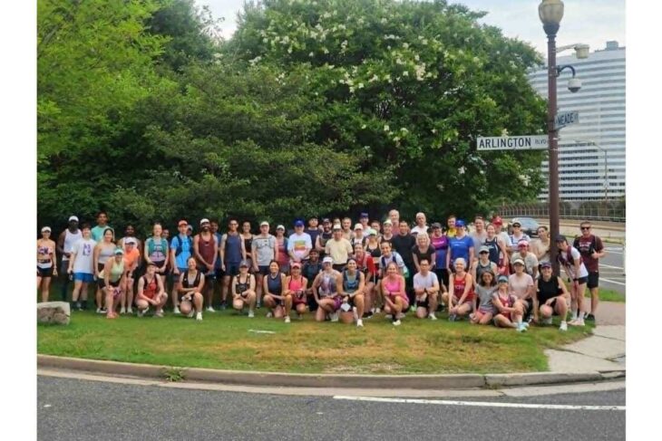 Large diverse group of people in athletic wear posing for a group photo on a grassy area near a street corner under a leafy tree.