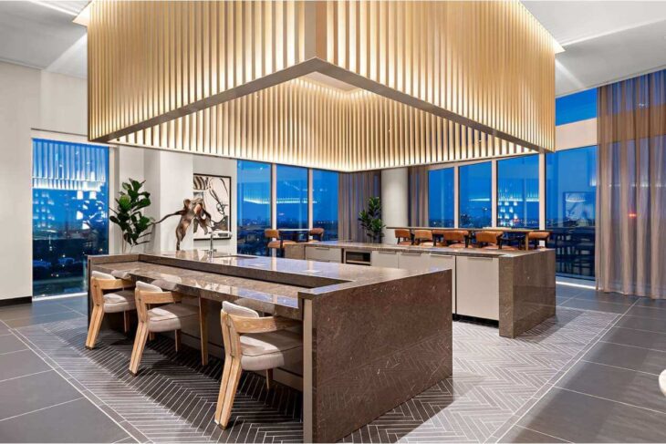 Open-plan lounge with a dark marble island, beige chairs, and a dramatic wooden grid ceiling; floor-to-ceiling windows overlook a city at night.