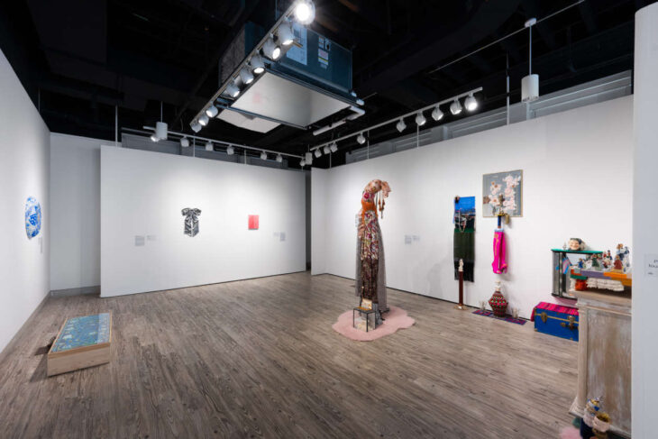 Wide view of a contemporary art gallery with white walls and wood floors, featuring a tall embellished dress sculpture center stage on a pink rug surrounded by other textile and mixed-media pieces on the right and left walls.