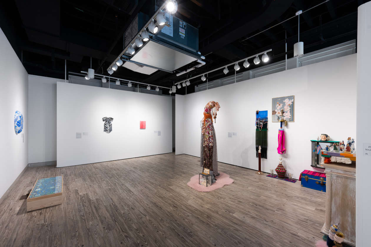 Wide view of a contemporary art gallery with white walls and wood floors, featuring a tall embellished dress sculpture center stage on a pink rug surrounded by other textile and mixed-media pieces on the right and left walls.