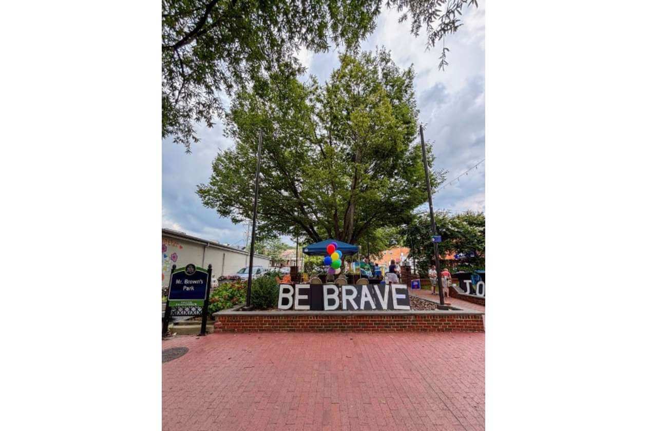 Park scene with a brick platform displaying BE BRAVE, colorful balloons, and a blue canopy under a large tree.