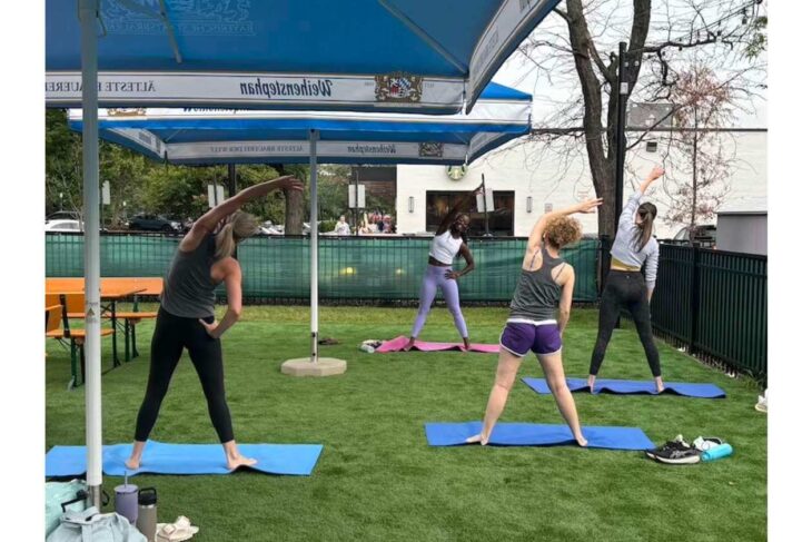 Group of women doing yoga stretches on blue mats under a blue canopy in a grassy outdoor area.