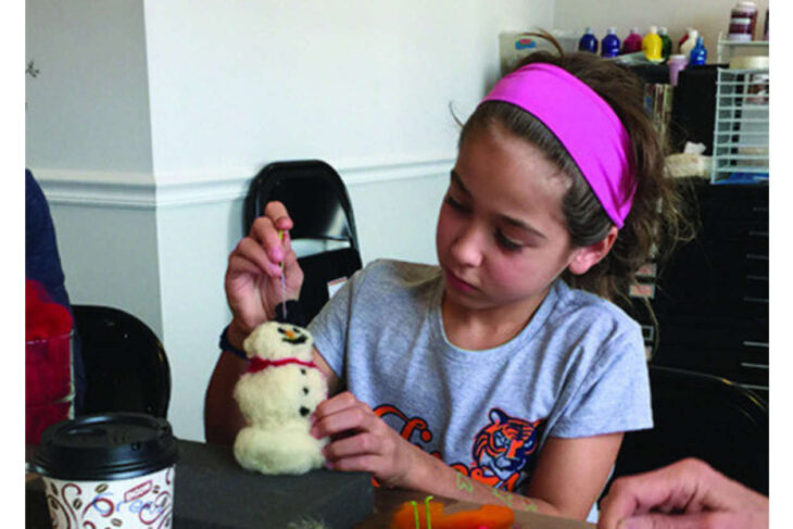 Girl with pink headband sewing a small plush snowman at a craft table.
