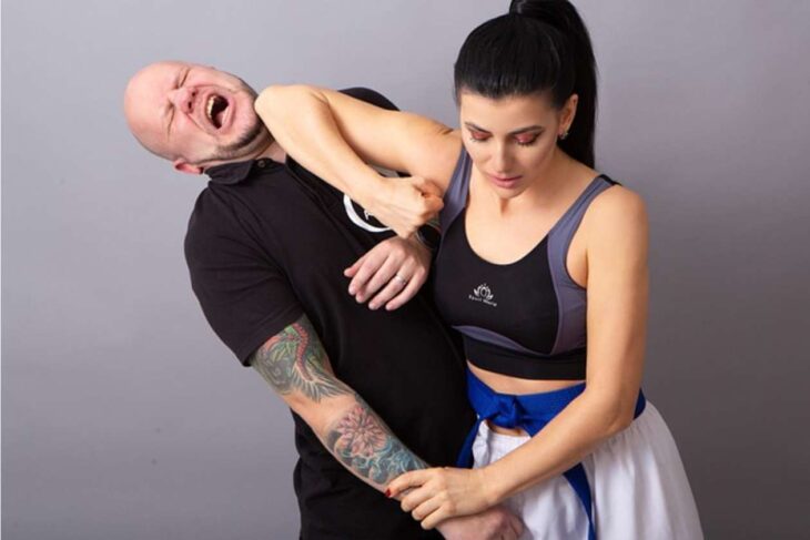 Self-defense demonstration: a woman applies an arm-bar to a man who grimaces in a gym-like setting.