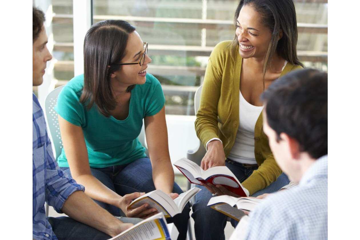 Group of young adults sitting together, reading books and smiling, likely studying or sharing a discussion in a casual setting