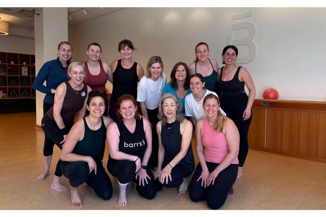Twelve smiling women in workout clothes posing together in a barre fitness studio.