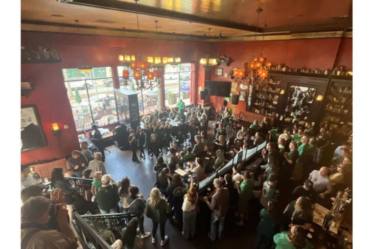 Crowded bar/restaurant interior with people standing and seated, wearing green, under warm chandeliers and red walls, near a performance area.