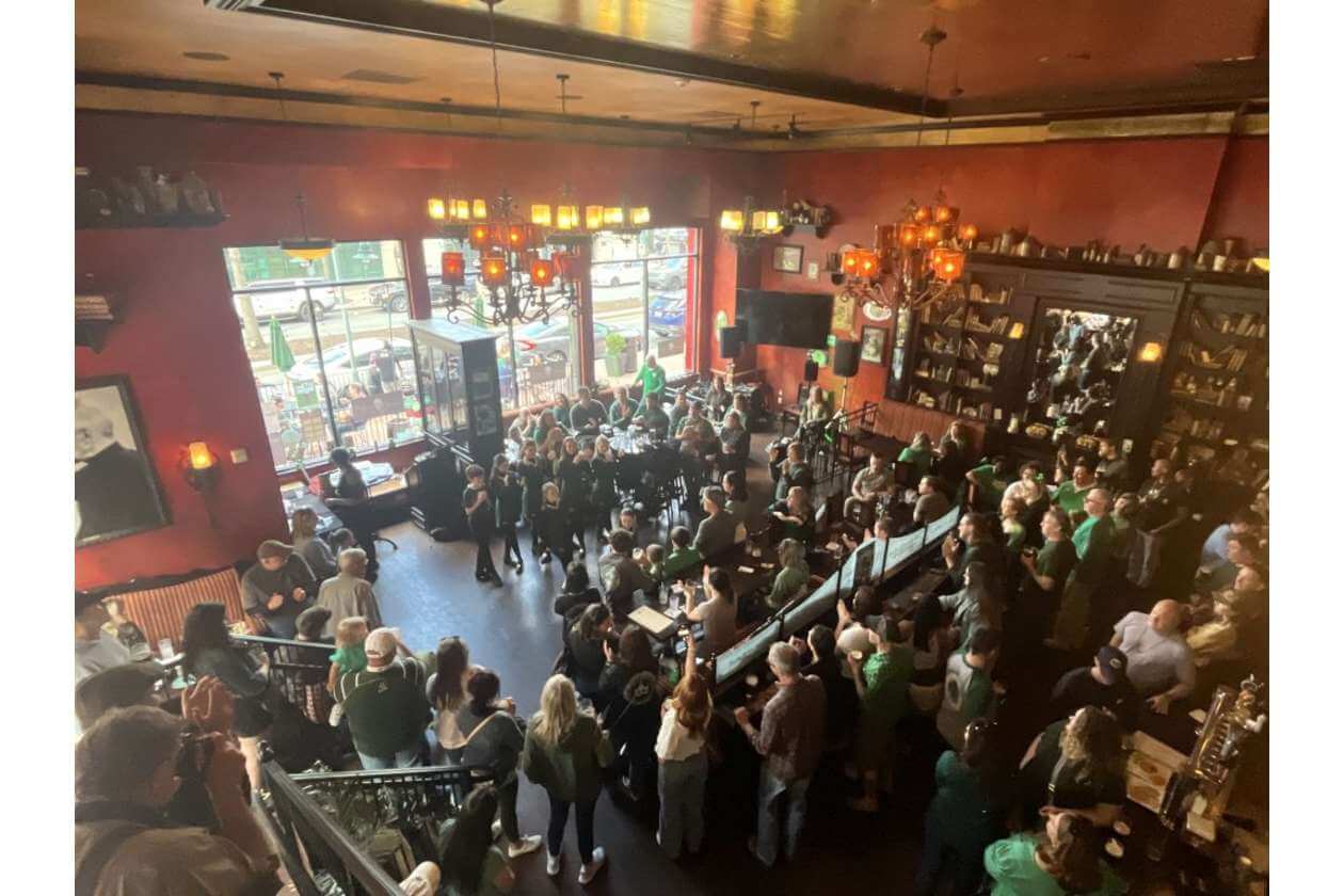 Crowded bar/restaurant interior with people standing and seated, wearing green, under warm chandeliers and red walls, near a performance area.