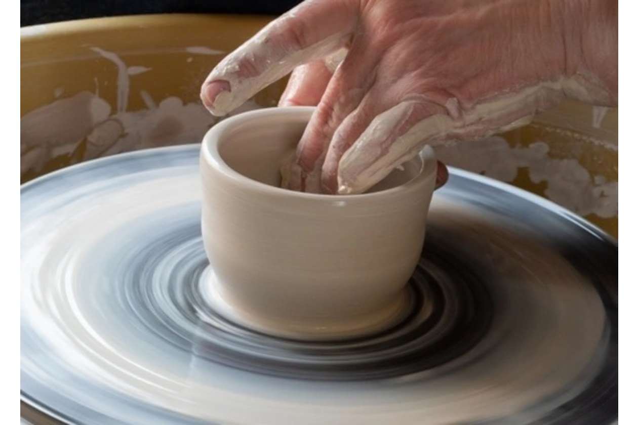 Hands shaping a wet clay mug on a spinning pottery wheel, forming the vessel's walls.