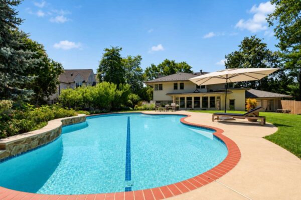 Rectangular curved pool with blue water, brick coping, and a shaded lounge area in a sunny backyard.