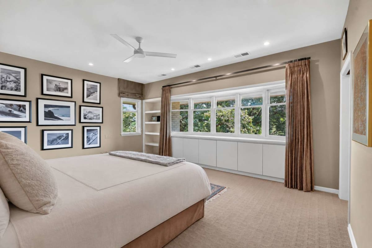 Bedroom with a beige wall gallery of black-framed photos, a large bed, and a row of windows with white cabinets beneath.