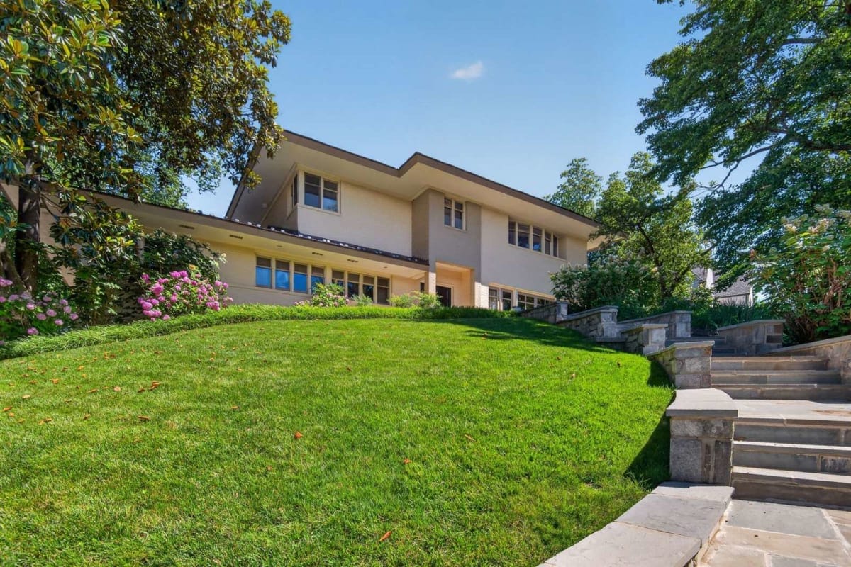 Modern two-story house atop a grassy hill with a stone staircase on the right and lush trees framing the scene.