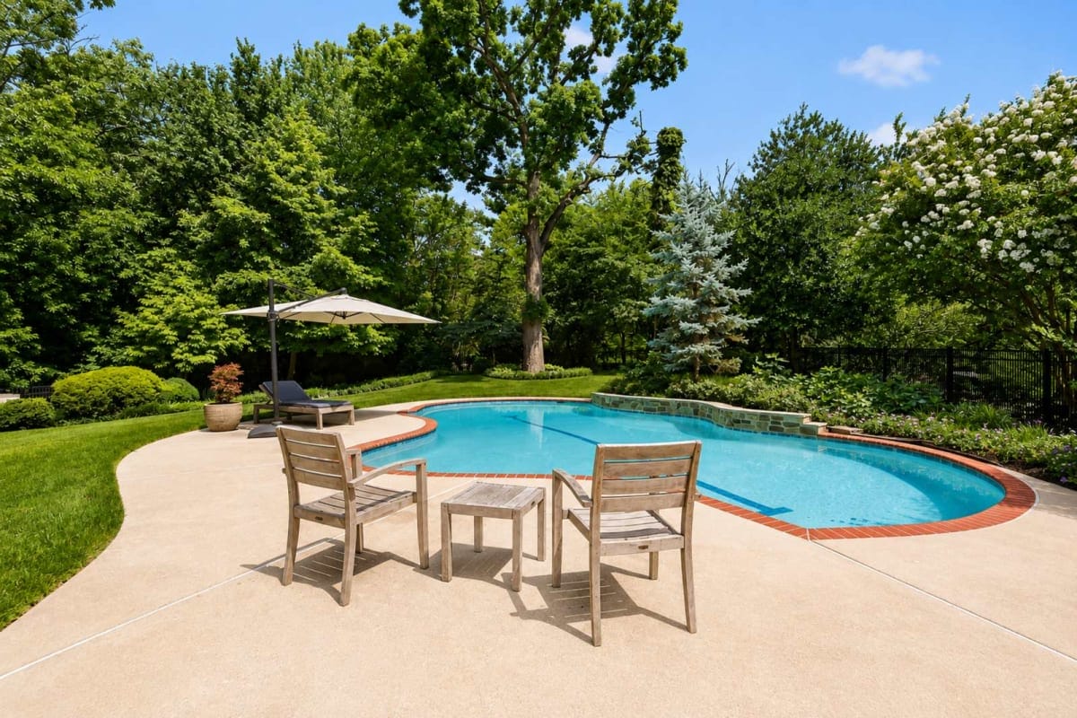 Backyard pool area with two wooden chairs and a small table on a concrete deck, a beige umbrella, and lush trees surrounding the space.