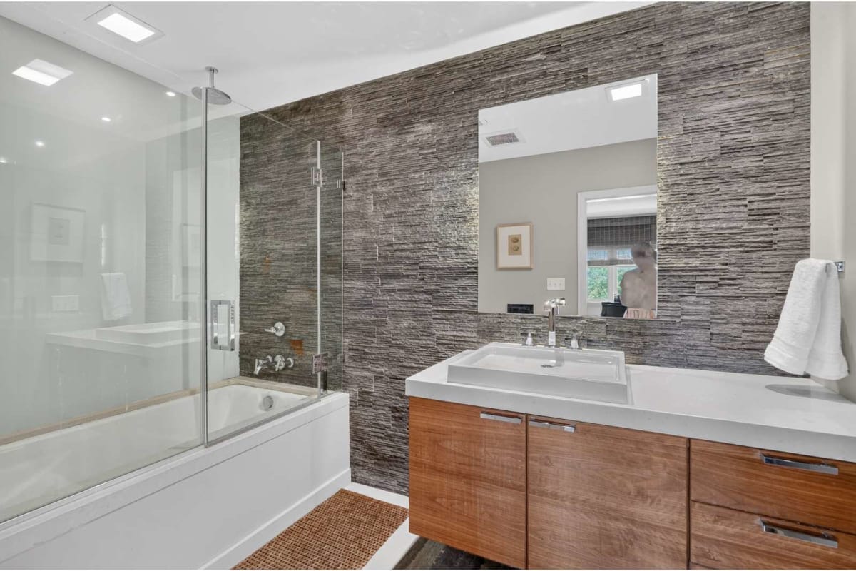 Modern bathroom with glass shower enclosure, white tub, and a wood-toned vanity under a large mirror.