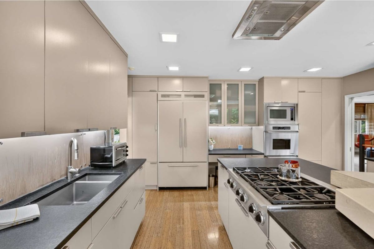 Modern beige kitchen with stainless steel appliances, large fridge, double ovens, and a central gas range island. Under-cabinet lighting and hardwood floors create a bright, contemporary space.