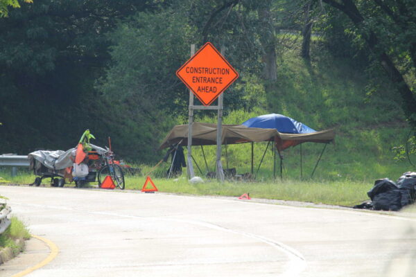 Orange diamond 'Construction Entrance Ahead' sign over a roadside campsite with a tarp shelter, bike, and gear