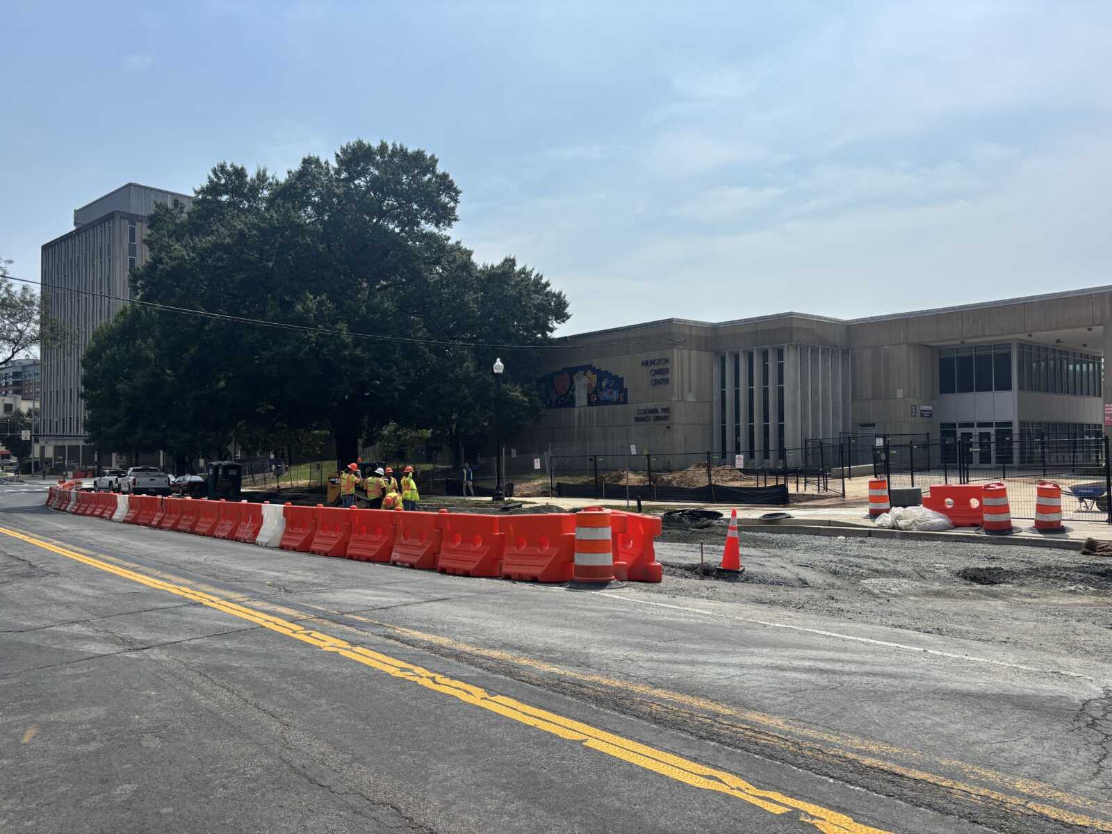 Construction workers in high-visibility vests stand near orange barriers blocking a street under renovation by a modern building in the background.