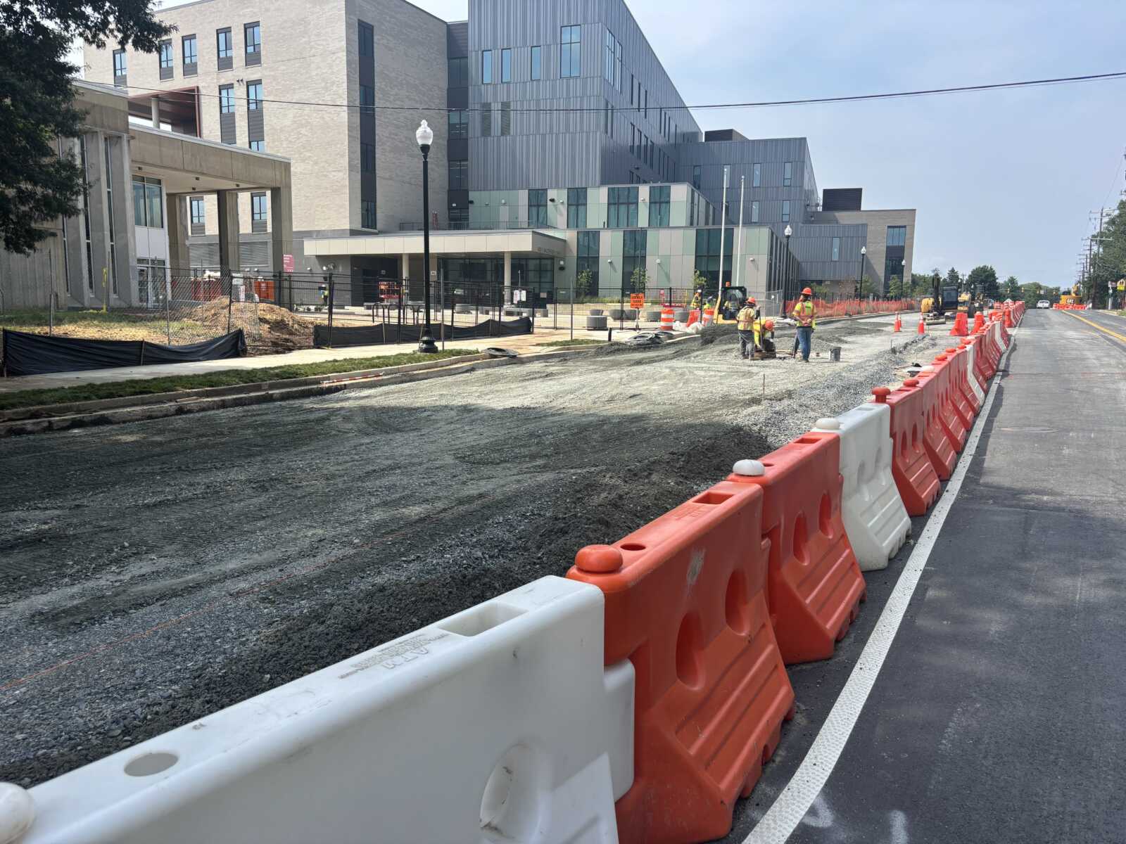 Street construction site with orange barriers, workers in safety vests, and a modern multi-story building in the background.