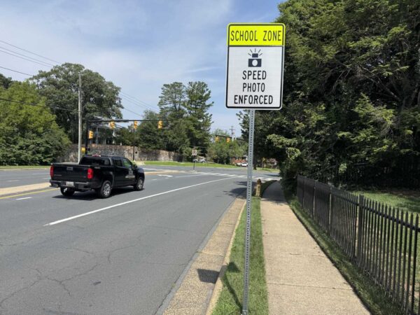 School zone sign with SPEED PHOTO ENFORCED above a street where a black pickup drives by, trees in the background