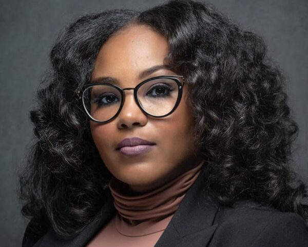 Professional headshot of a woman with curly black hair, round glasses, brown turtleneck, and blazer against a gray backdrop.