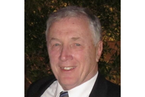 Smiling older man with gray hair in a dark suit and striped tie, outdoors at night with greenery in the background.