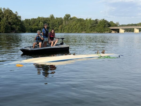 Three people wearing life jackets sit on a small motorboat on a calm lake, with a capsized white kayak nearby and a forested shoreline in the background.