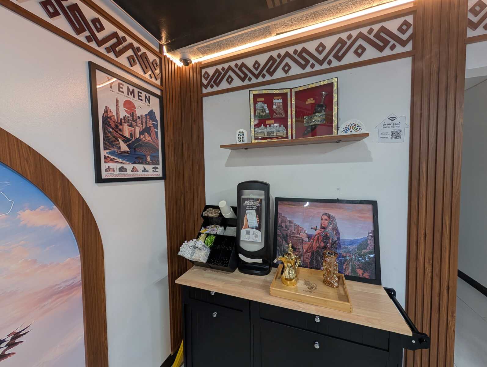 Travel-themed interior corner featuring a Yemen poster, wood trim lattice, and framed city prints above a wooden shelf.